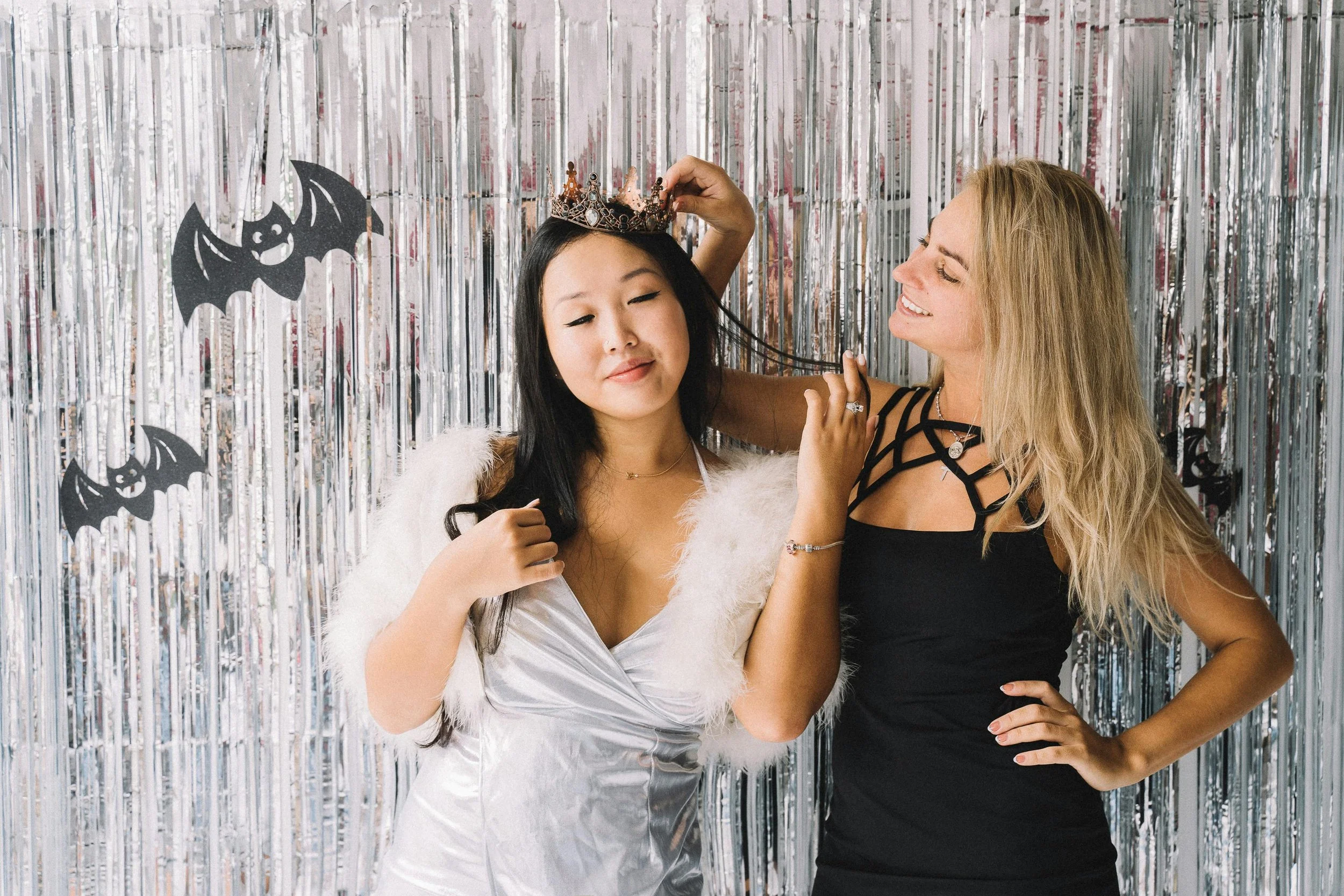 Two women at a Halloween party with a silver tinsel backdrop and bat decorations. One woman is wearing a white dress with a fur stole and a crown, while the other woman is in a black dress.