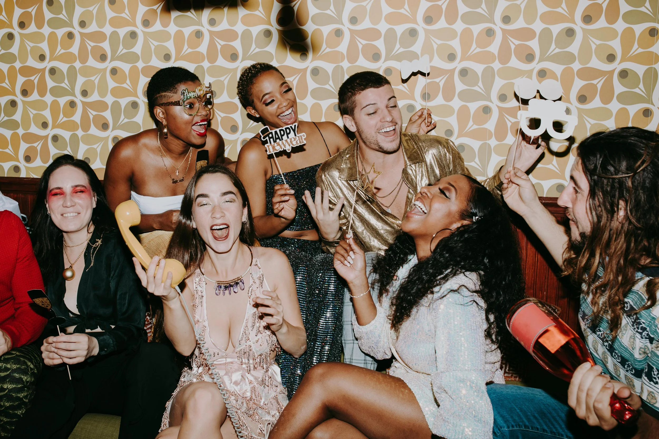 Group of friends celebrating a party with playful props, laughing and smiling in a cozy setting, with colorful wallpaper in the background.