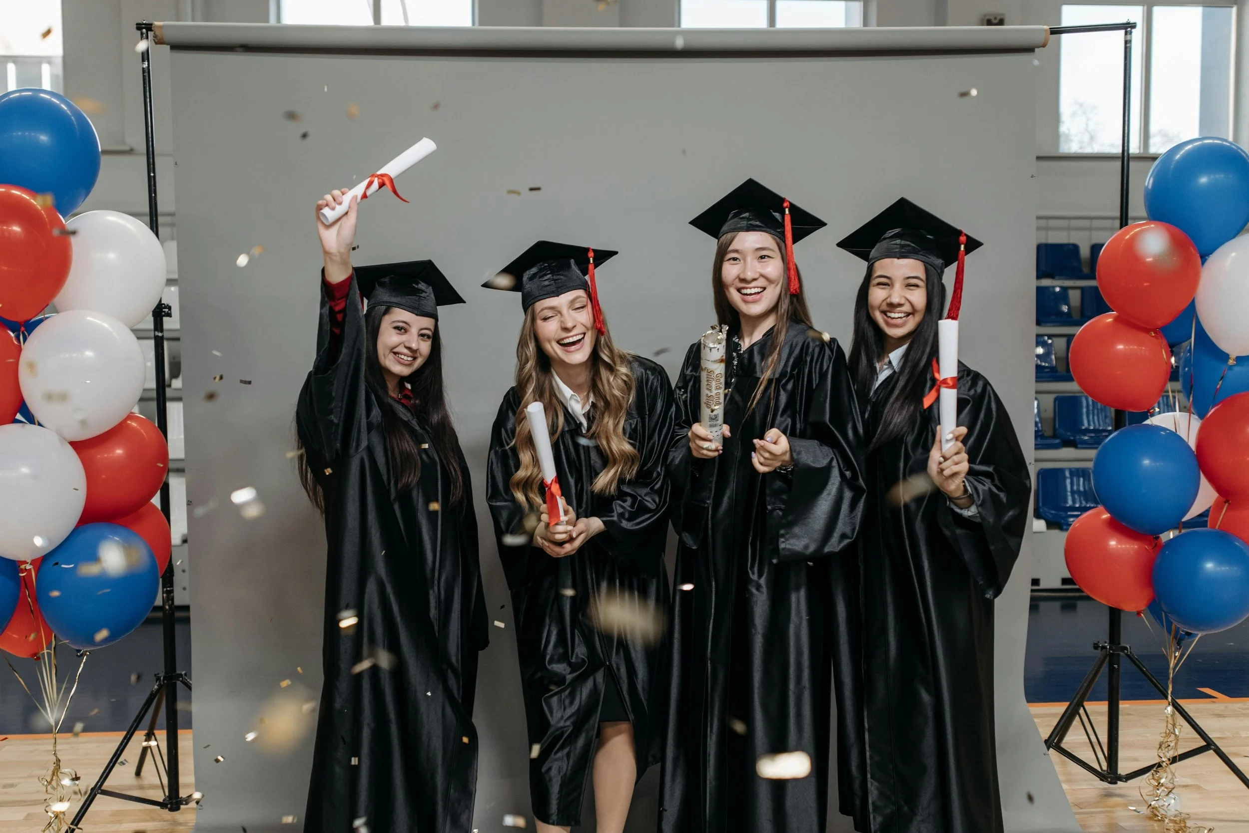 Four female graduates in black caps and gowns celebrating with diplomas and a scroll, standing in front of a gray backdrop, surrounded by red, white, and blue balloons, with confetti falling around them.