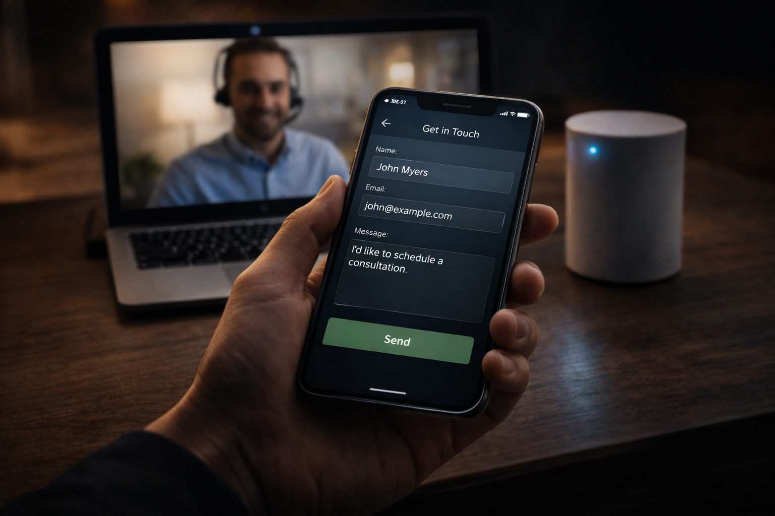 Person holding smartphone with a contact form on the screen, in front of a laptop showing a man wearing headphones, and a white smart speaker on a wooden table.