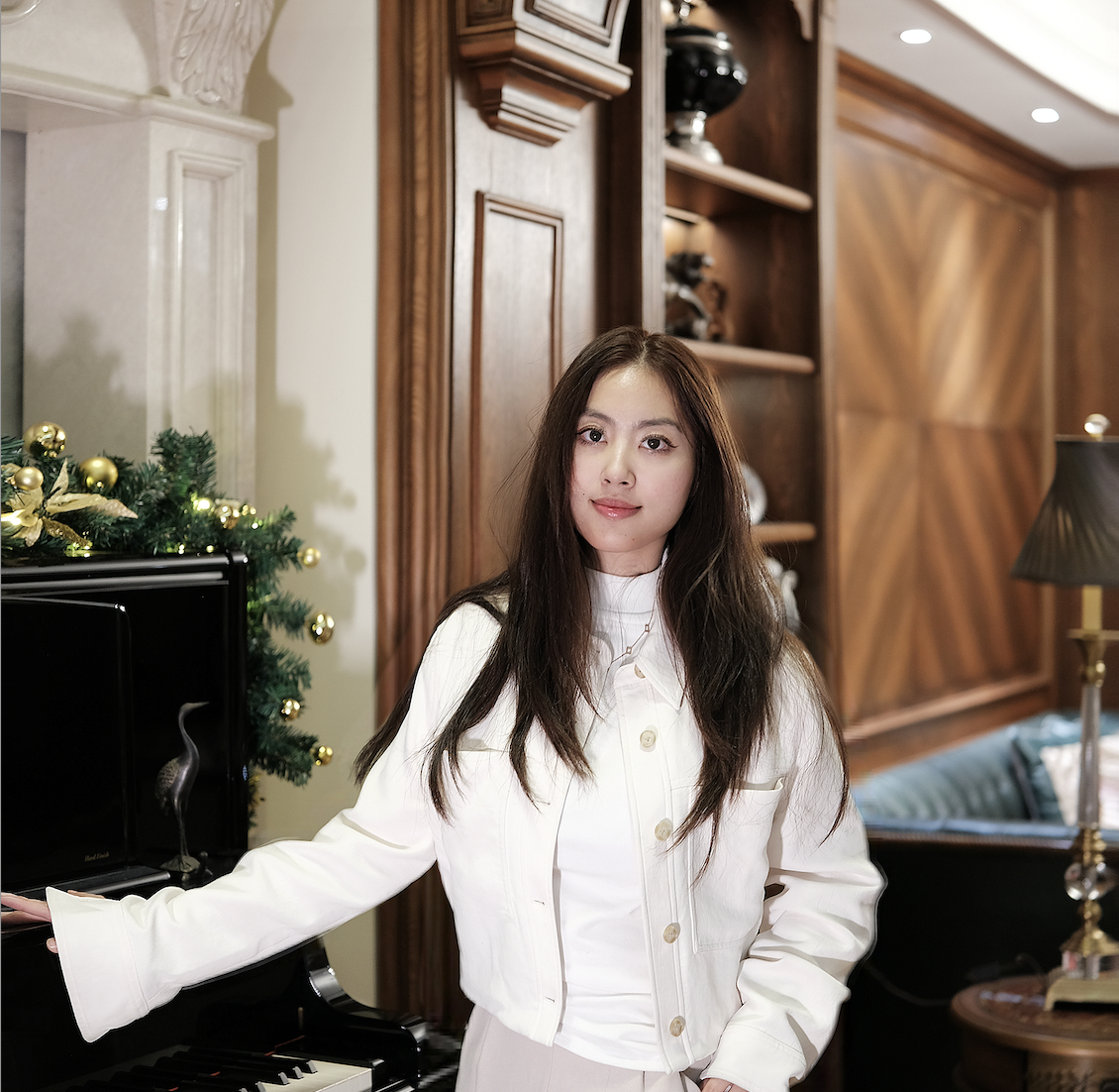 A young woman with long dark hair standing near a black piano with Christmas decorations, in a warmly lit room with wooden wall paneling and shelves.
