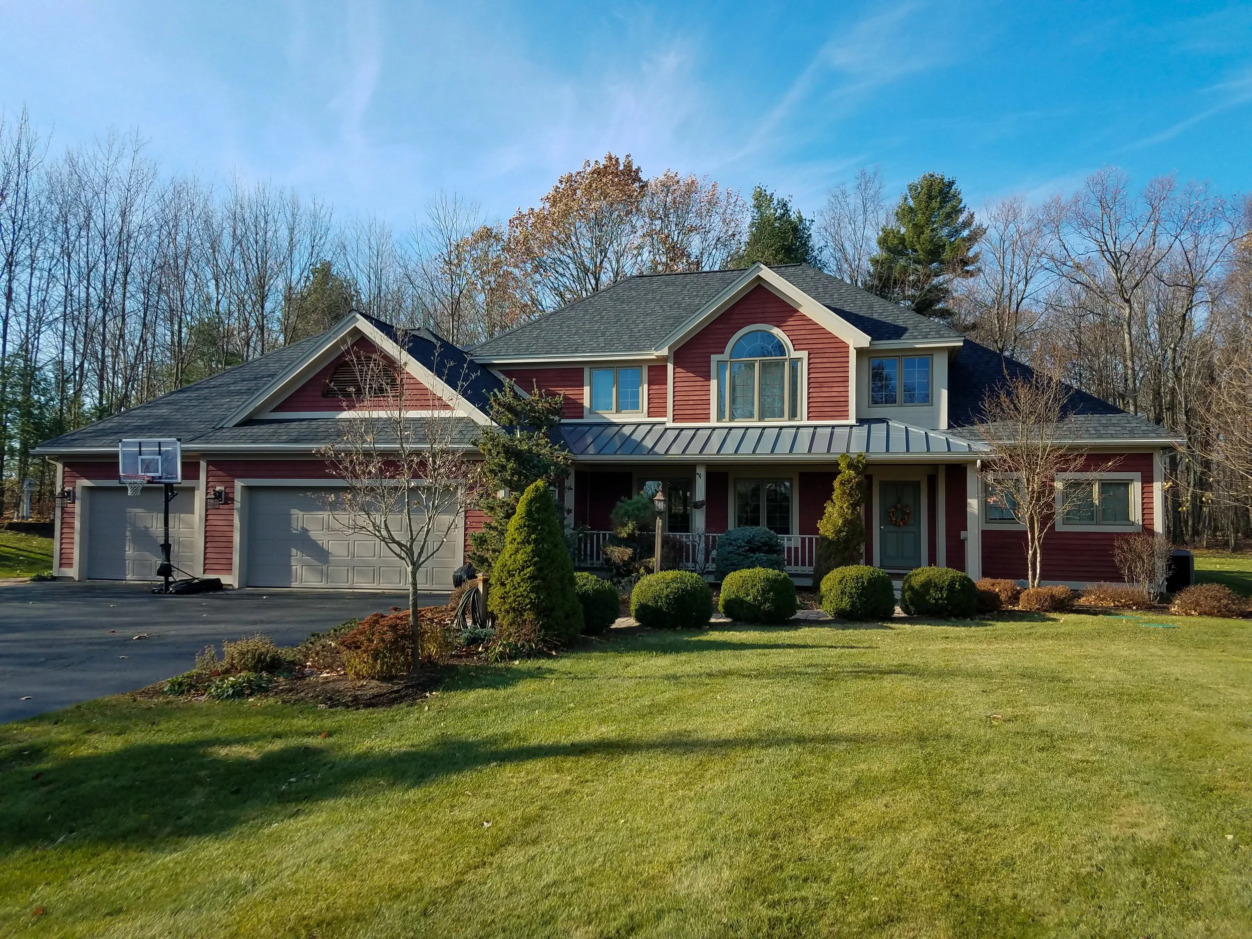 A two-story red house with white trim and a gray roof, surrounded by a well-maintained lawn and shrubs, with some trees in the background and a blue sky overhead.