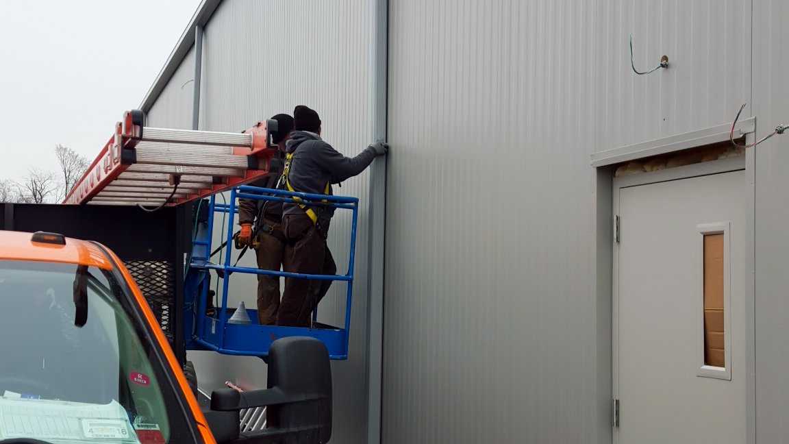 Two workers installing or repairing metal panels on the exterior of a building, using a lift platform.
