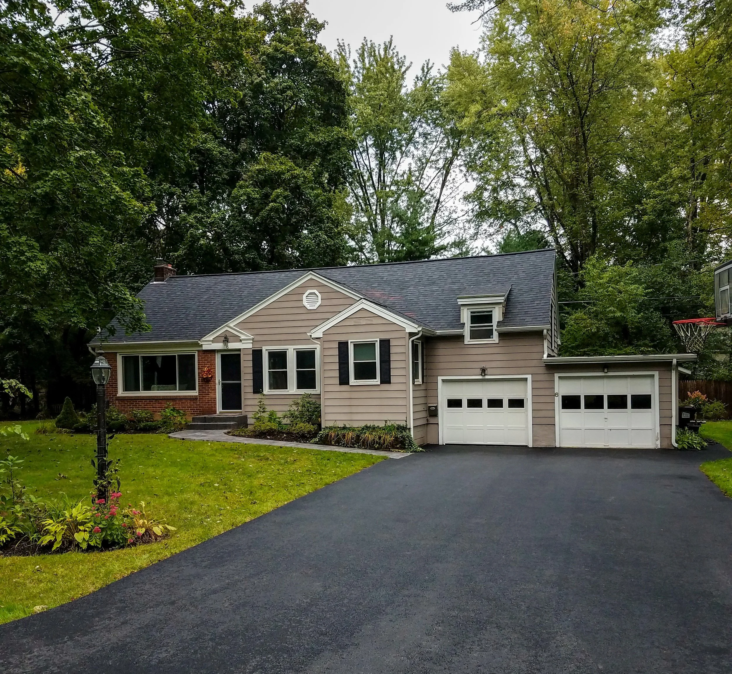 Front view of a suburban house with beige and brick exterior, black shutters, three-car garage, asphalt driveway, and lush green lawn with flower beds and trees in the background.