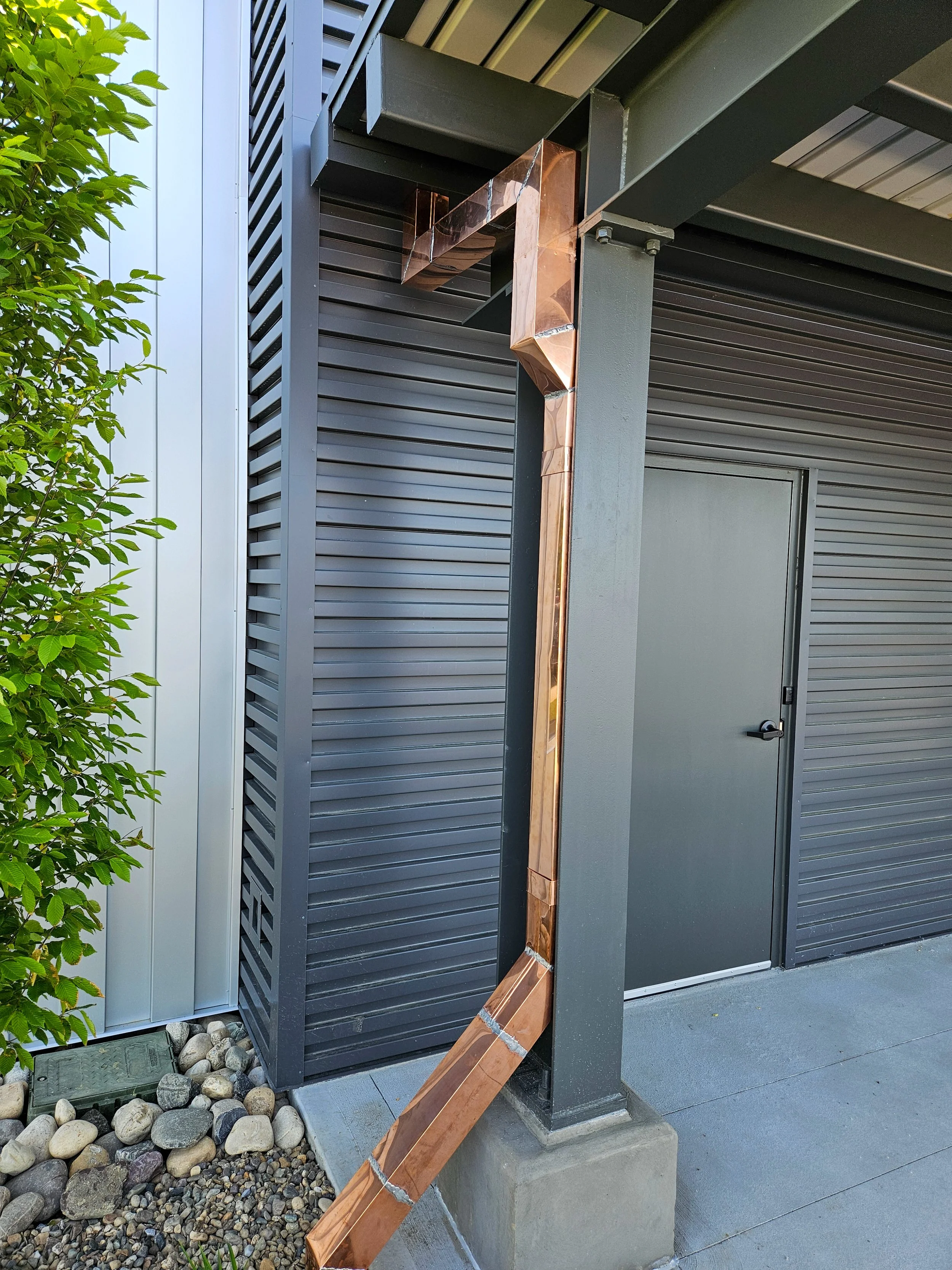 Copper downspout attached to the corner of a building with gray siding, next to a closed gray door and some landscaping rocks.
