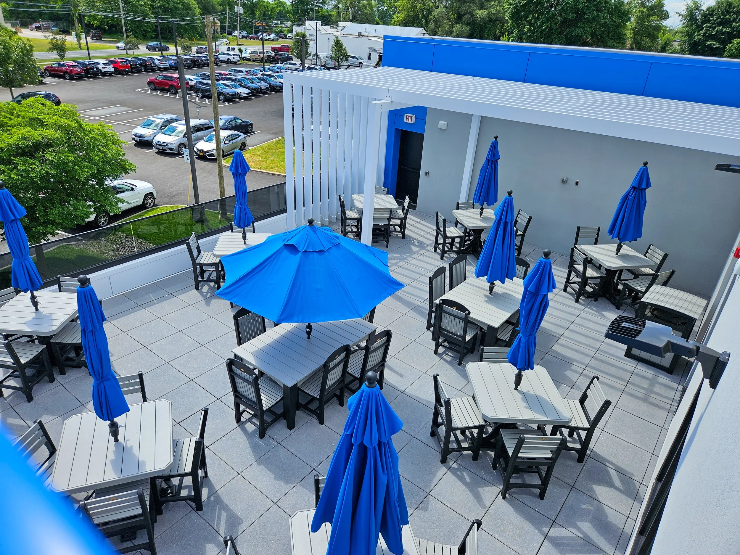 Empty outdoor patio with tables, chairs, and blue umbrellas on a sunny day, overlooking a parking lot and green trees.