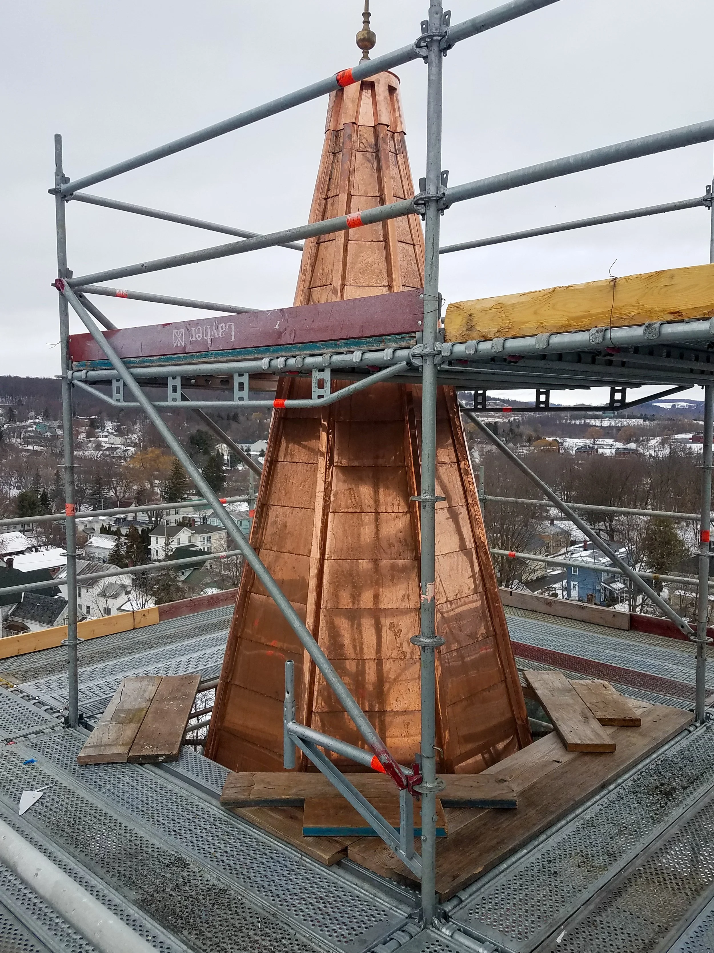 Copper-roofed spire on a building under construction, surrounded by scaffolding, against a cloudy sky and a distant snowy town.