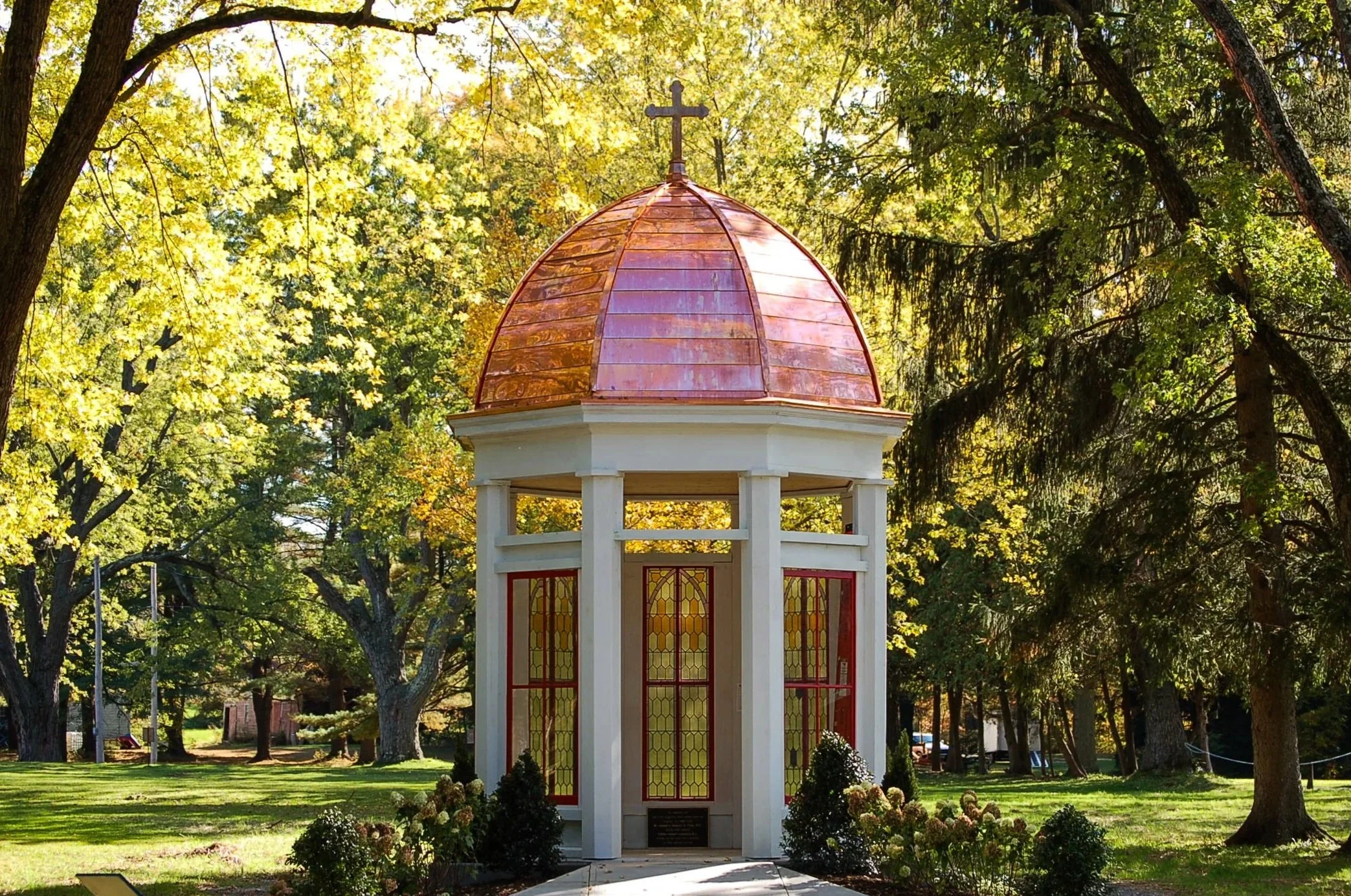 Small white pavilion with a copper dome and stained glass windows, surrounded by trees and greenery in a park.