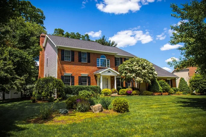 Large brick house with a manicured lawn, trees, and shrubs on a sunny day with a blue sky and scattered clouds.