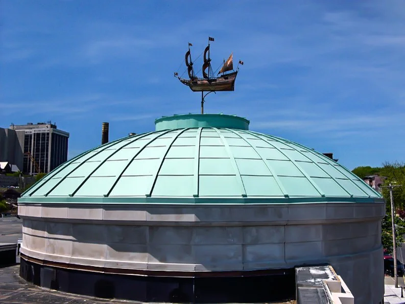 A rooftop green dome with a decorative weather vane shaped like a sailing ship on top, against a clear blue sky.