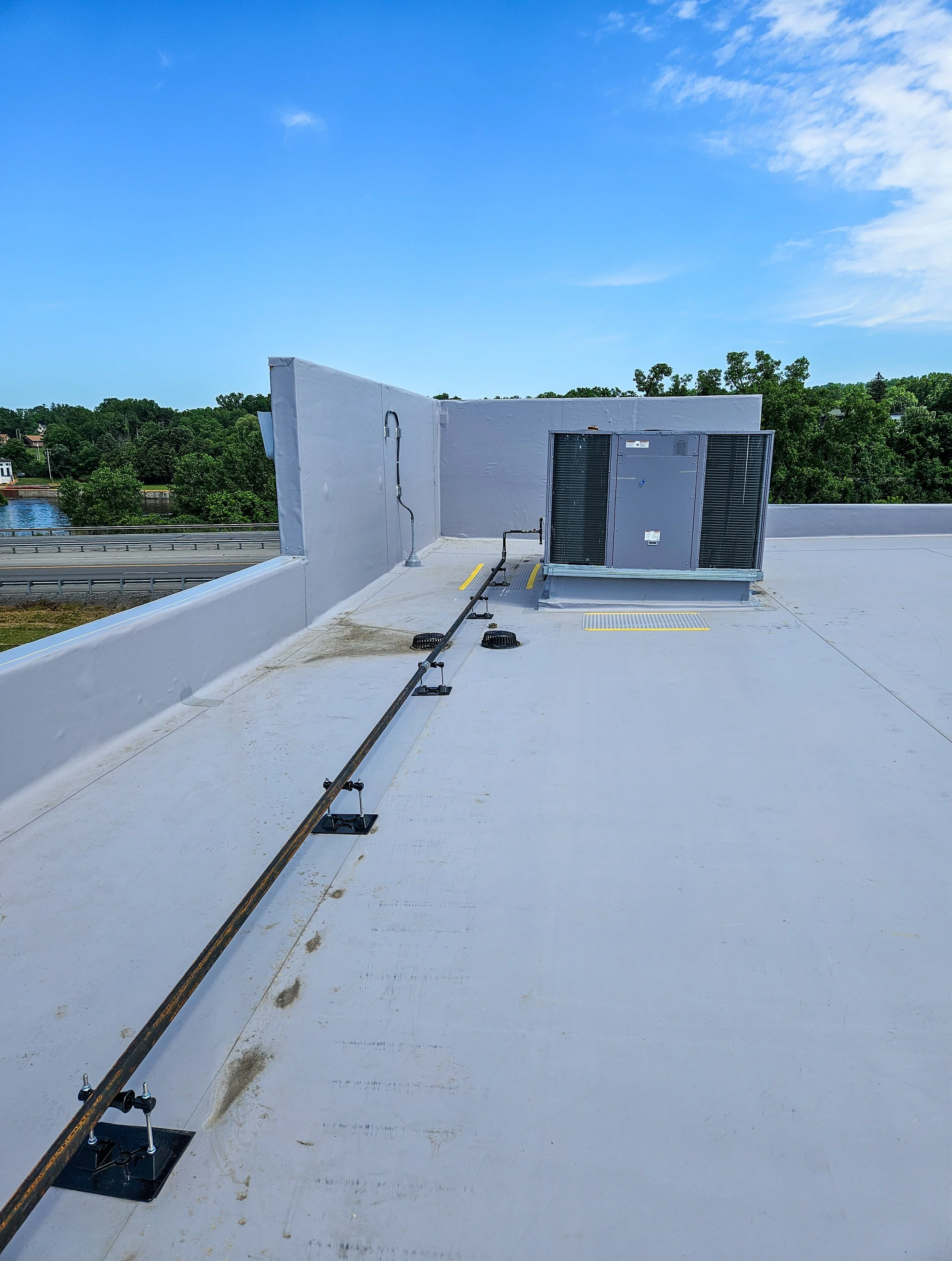 View of a flat rooftop with an HVAC unit, electrical piping, and a railing, against a backdrop of green trees and a blue sky.