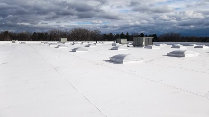 Overhead view of a flat white commercial building roof with multiple HVAC units and vents, under a cloudy sky.