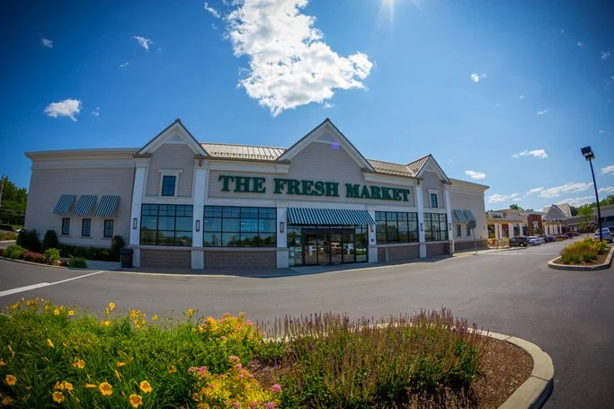 Exterior of a grocery store named 'The Fresh Market' with a parking lot and landscaping in front, under a blue sky with clouds.