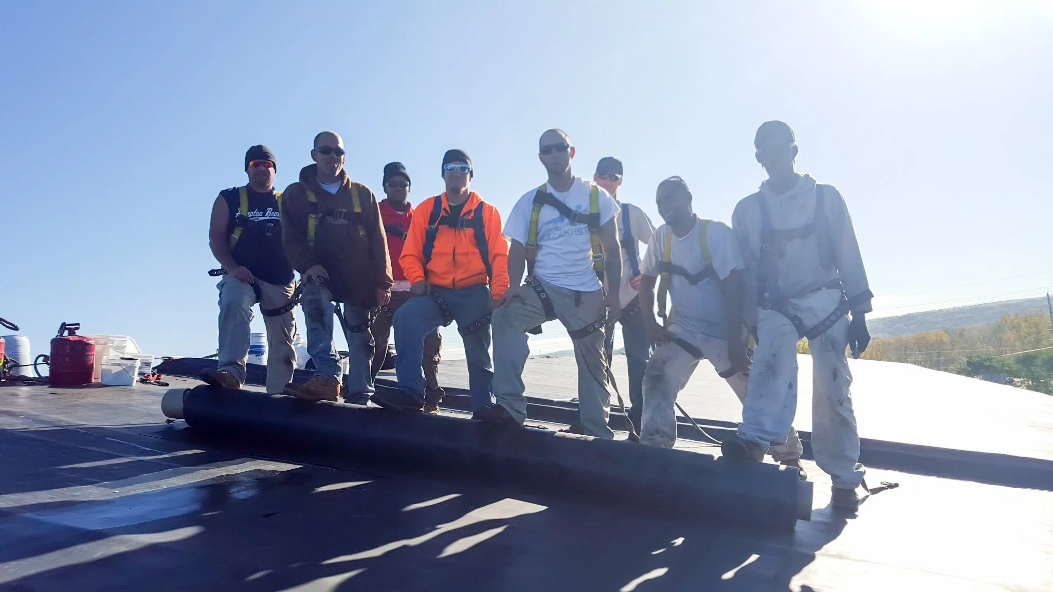 Group of nine people standing on a roof with construction equipment, wearing harnesses and outdoor clothing, under a clear sky.