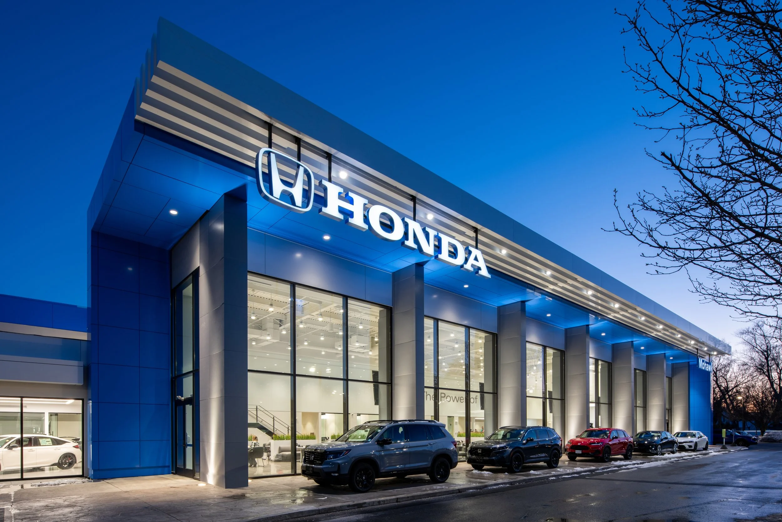 Exterior of a Honda car dealership building during dusk, with large glass windows and cars parked outside, illuminated by blue exterior lighting.