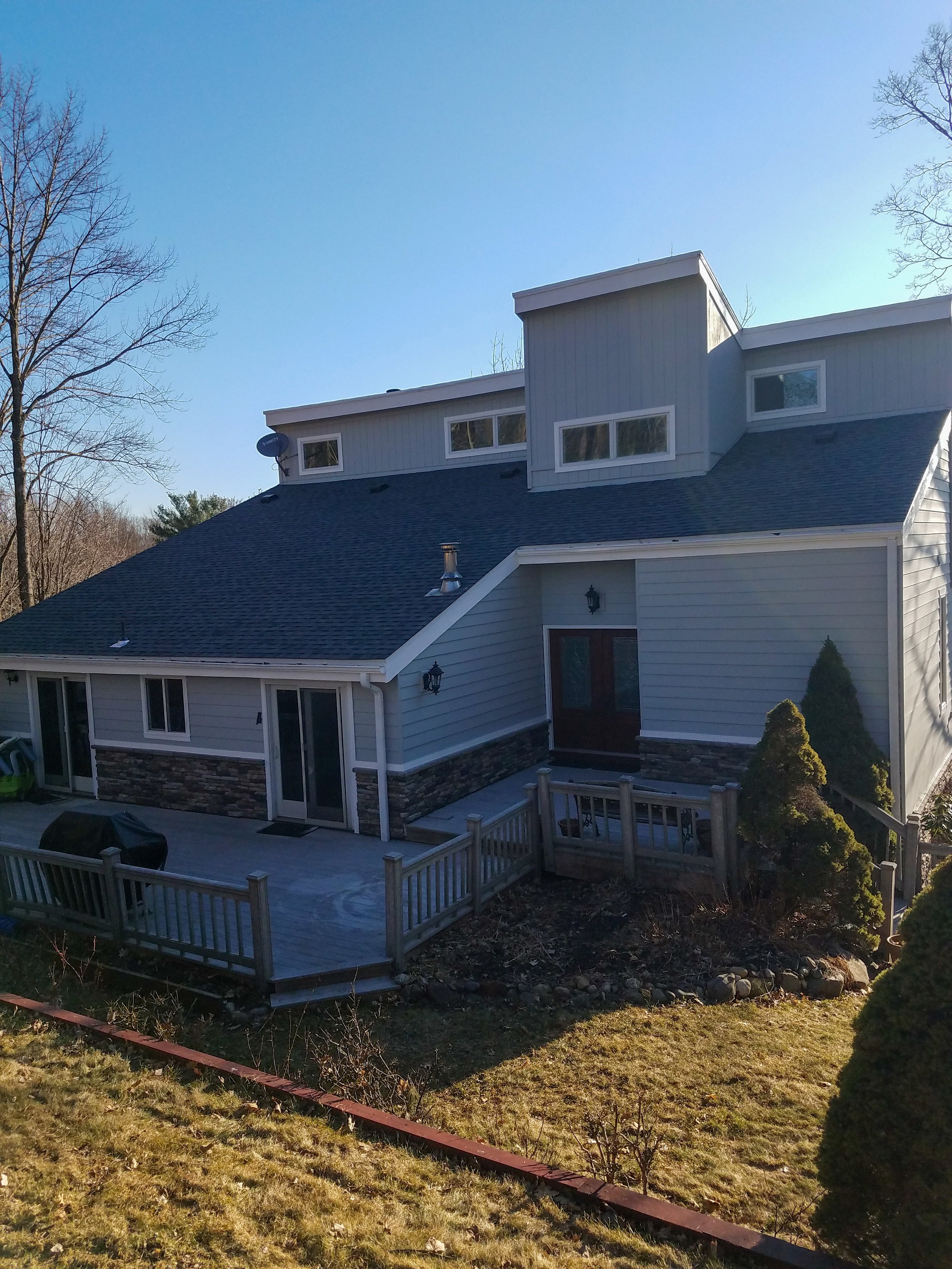 A modern two-story house with a dark shingle roof, light gray siding, and stone accents. It has a backyard with a wooden deck, a barbecue grill, and some landscaping with bushes and a grassy area. The sky is clear and the trees in the background are 