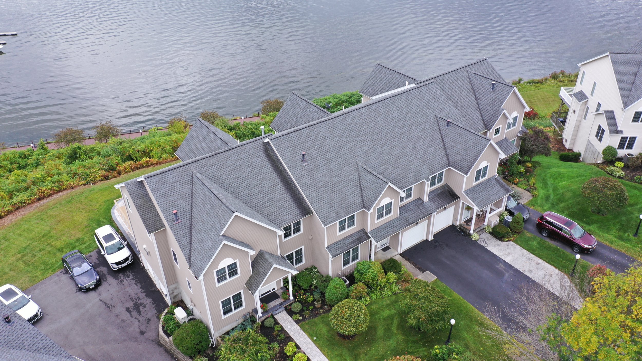 Aerial view of a multi-family residential complex with gray roofs and well-maintained green yards, located near a body of water, with parked cars in the driveways.