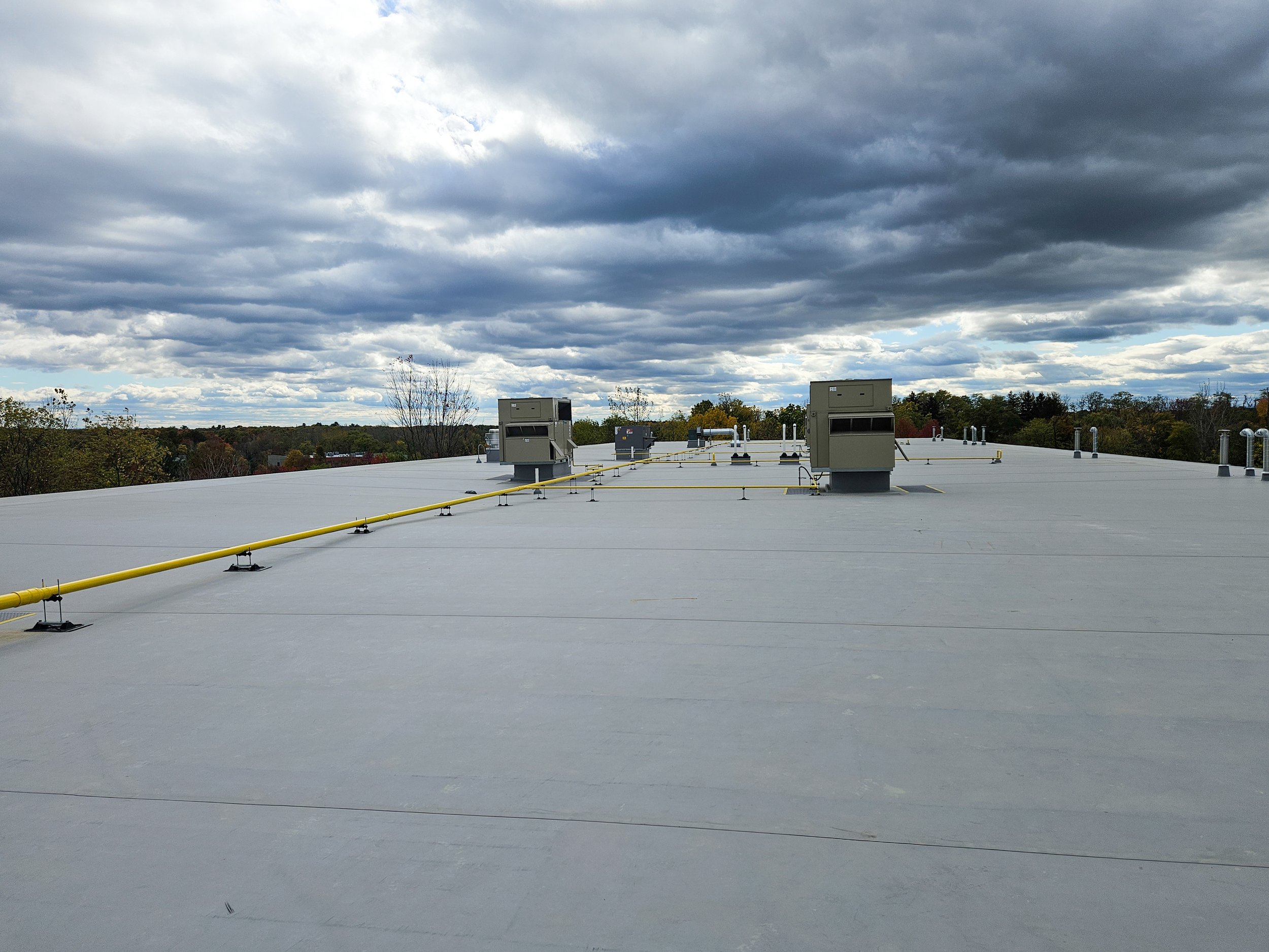 A flat rooftop with grey surface and multiple HVAC units, pipes, and vents under a cloudy sky.