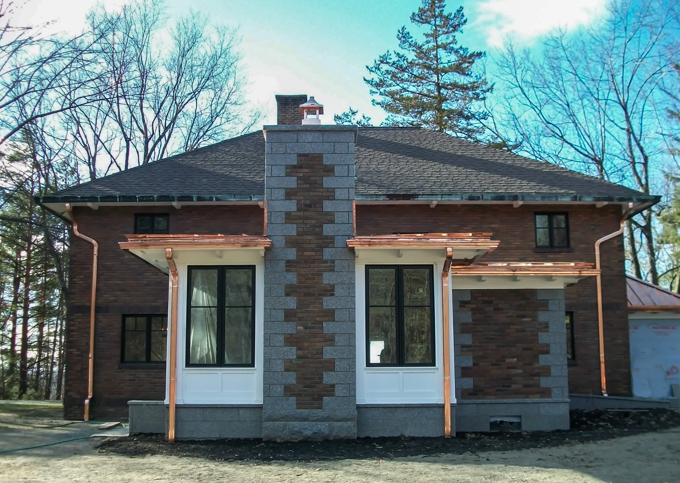 Front view of a partially constructed brick house with a chimney, large windows, and copper gutters, surrounded by trees.