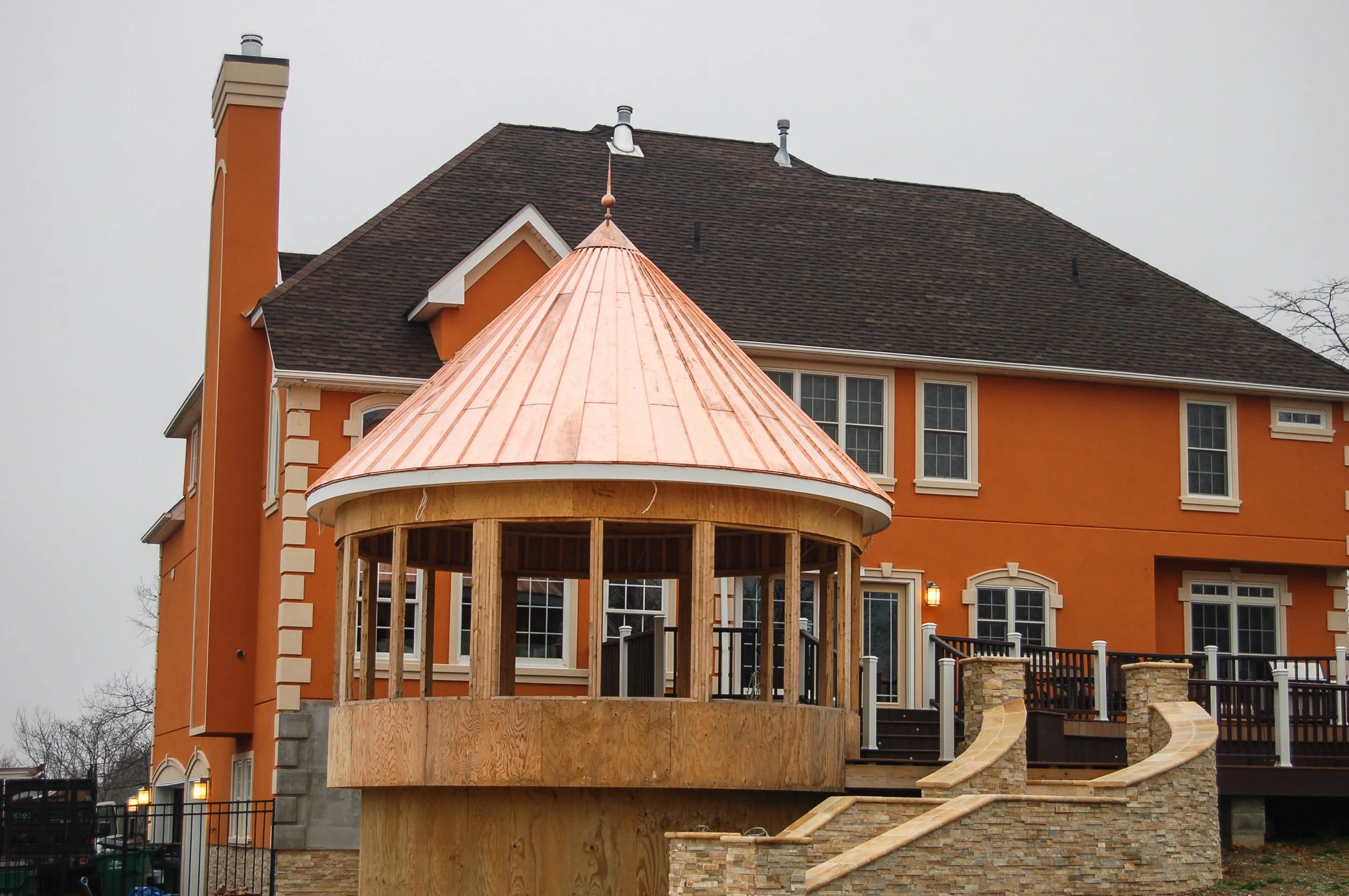 Under construction small round wooden structure with a copper roof in front of a large orange house with dormer windows and a sloped roof.