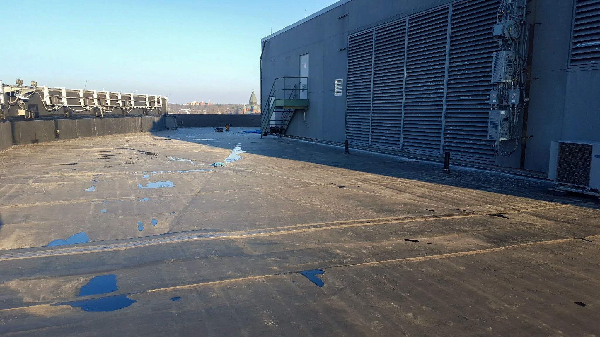 Rooftop view showing a flat surface with patches of water, a staircase, and equipment mounted on the building wall with a city skyline in the background.