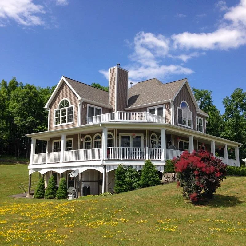 Large beige house with multiple levels, screened porch, and decorative arches, surrounded by green lawn and trees, under a blue sky with clouds.
