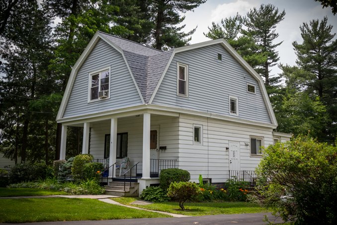 A two-story house with a distinctive curved roof, white siding, and a front porch with steps leading up to it, surrounded by greenery and trees.