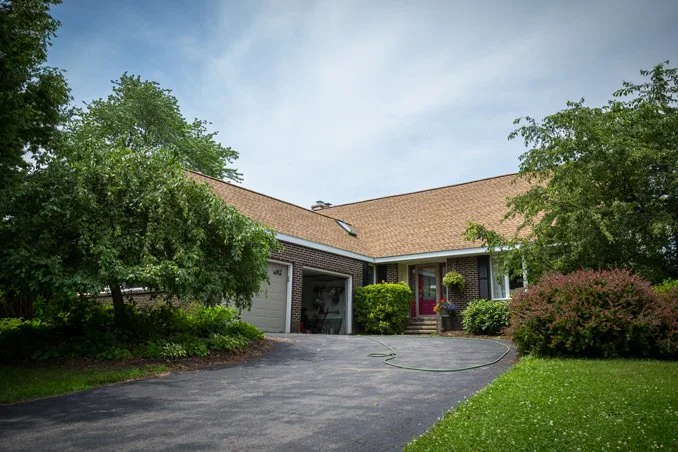 Front view of a suburban house with a driveway, garage, and front yard with bushes and trees under a cloudy sky.