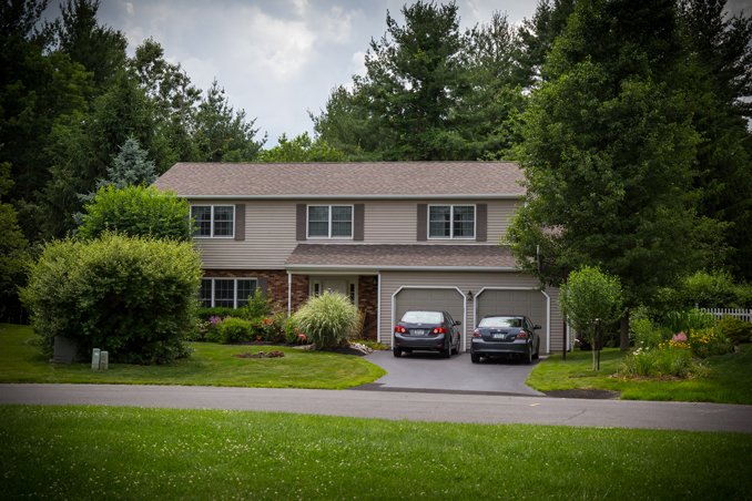 A two-story suburban house with a driveway, two parked cars, surrounded by green trees and shrubs.