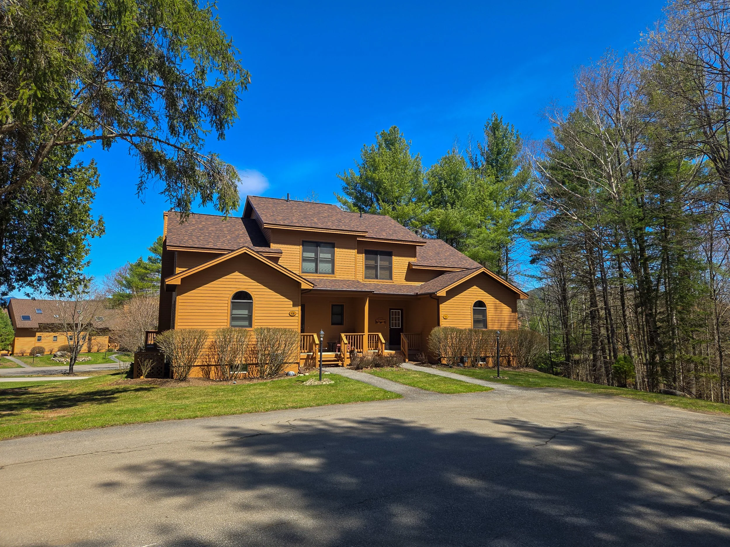 A two-story house painted in shades of brown with a gabled roof, surrounded by green lawns and trees, under a bright blue sky.