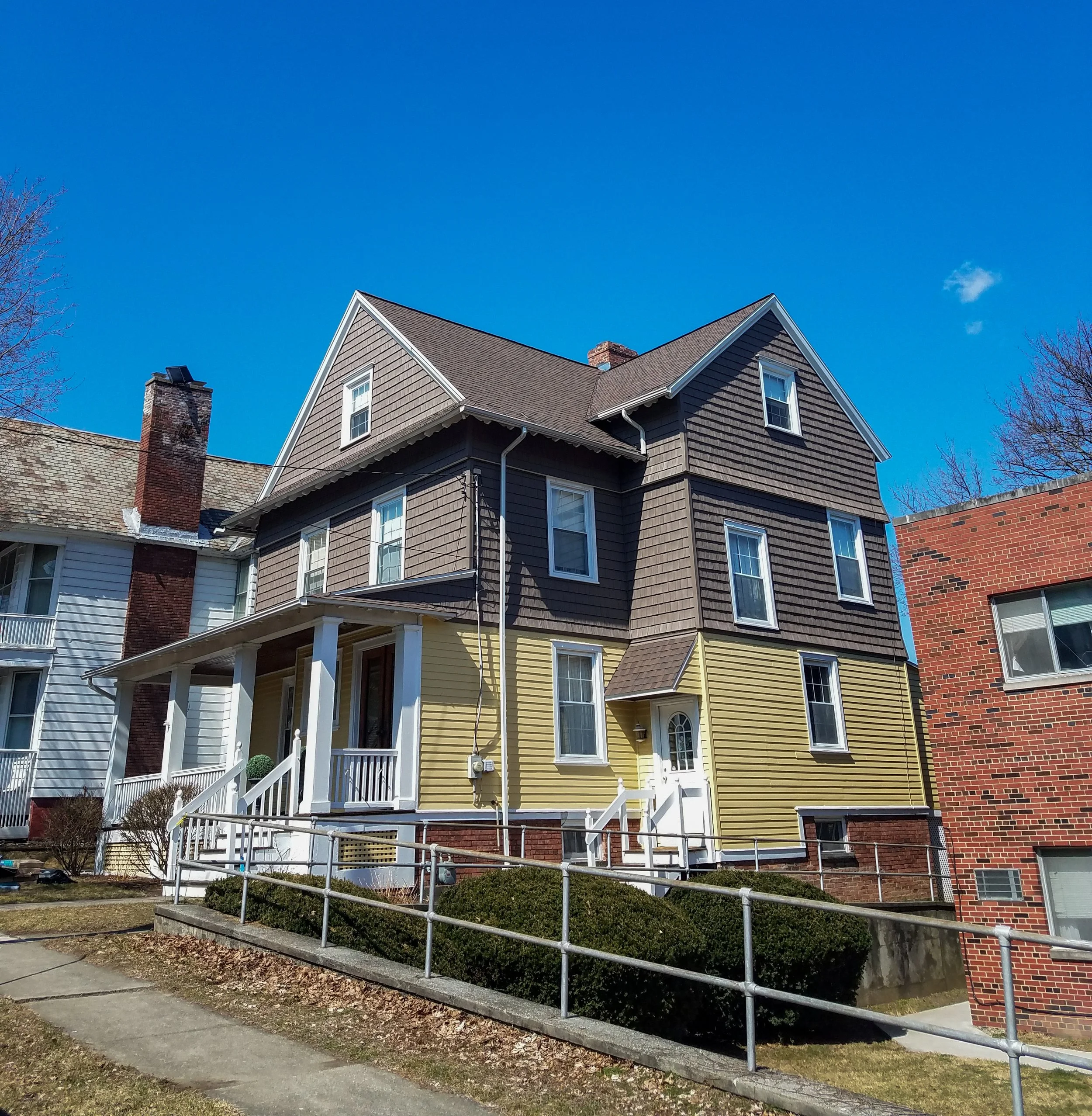Colorful multi-story house with brown, yellow, and darker brown exterior siding, front porch, and a walk-up ramp, situated on a sidewalk with bushes under a clear sunny sky.