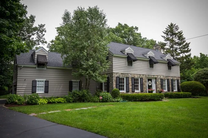 Residential house with gray siding, black shutters, stone accents, multiple dormer windows, surrounded by green lawn and landscaping, during overcast weather.