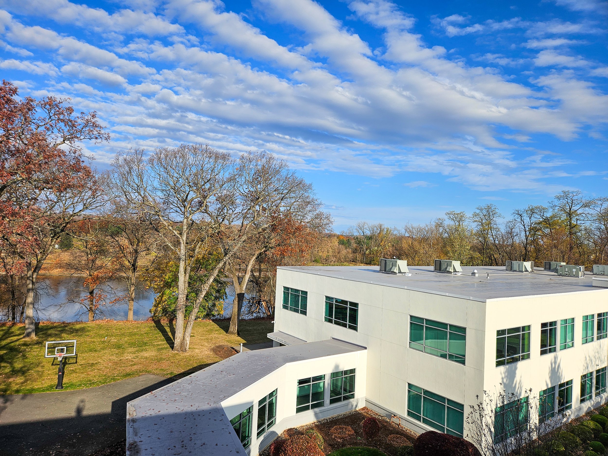 A modern white building with large windows, surrounded by trees with fall foliage, near a lake and a basketball hoop, under a partly cloudy sky.