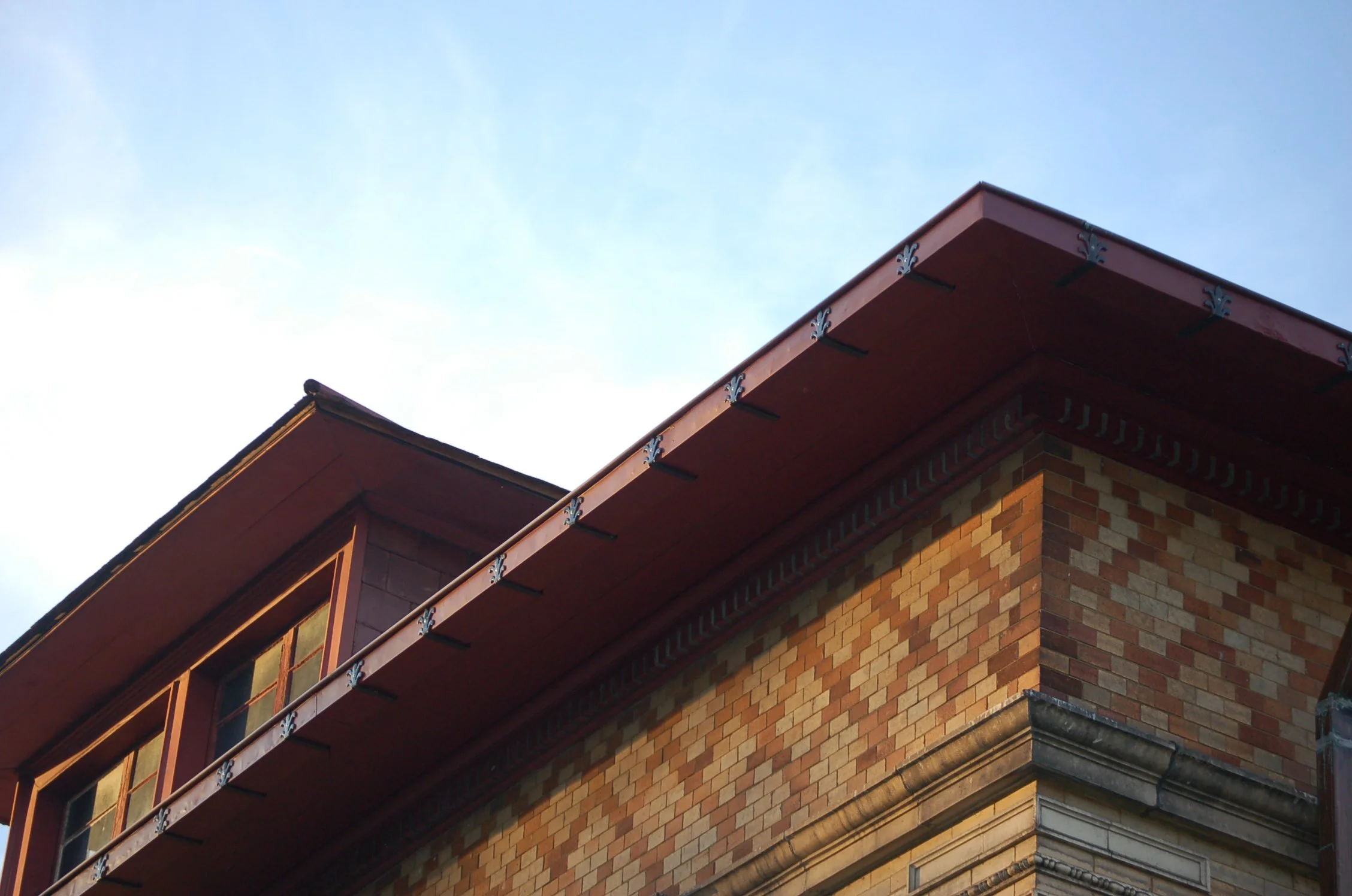 Close-up of a brick building corner with a metal roof edge and decorative brackets, under a partly cloudy sky.