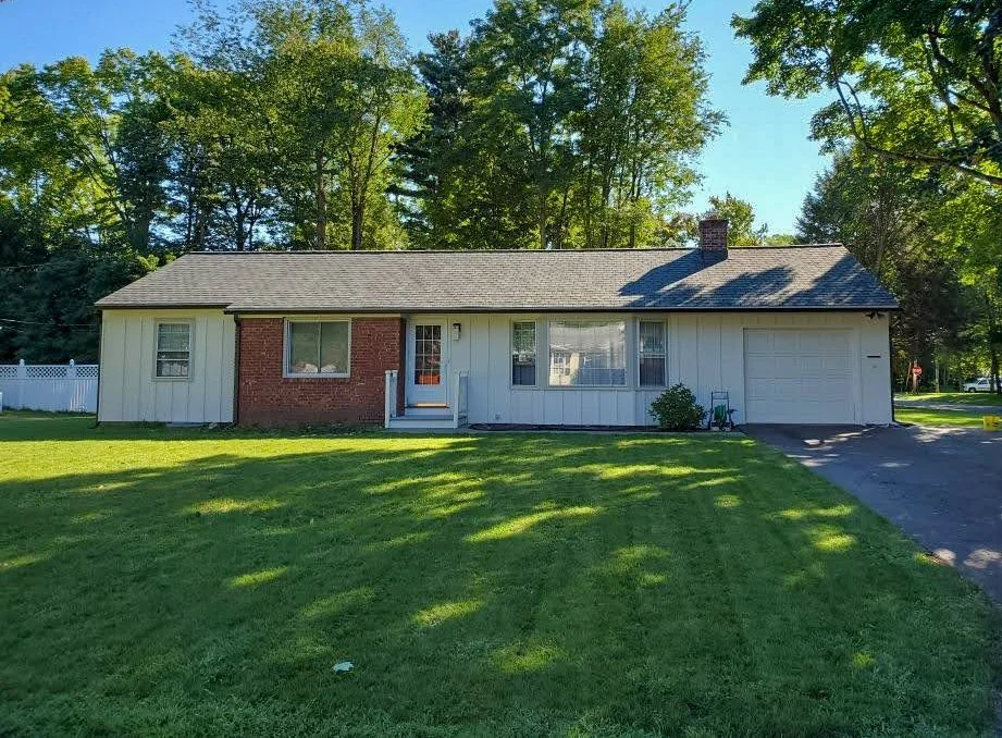 A single-story house with a brick and white siding exterior, a gray asphalt shingle roof, a small front porch with steps, and a one-car garage on the right. The house is surrounded by a green lawn and trees in the background.