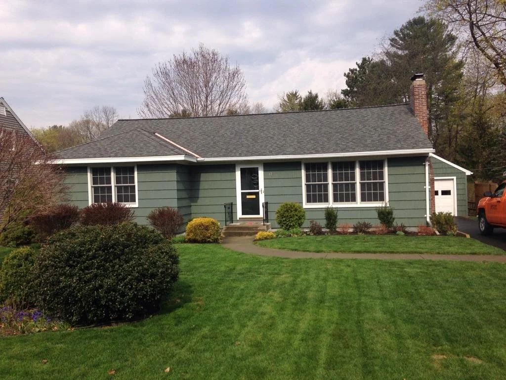 A single-story, green house with white trim, large front windows, and a driveway on the right side with an orange vehicle parked. The house has a black front door with a small staircase and railing. There is a landscaped front yard with green grass, 