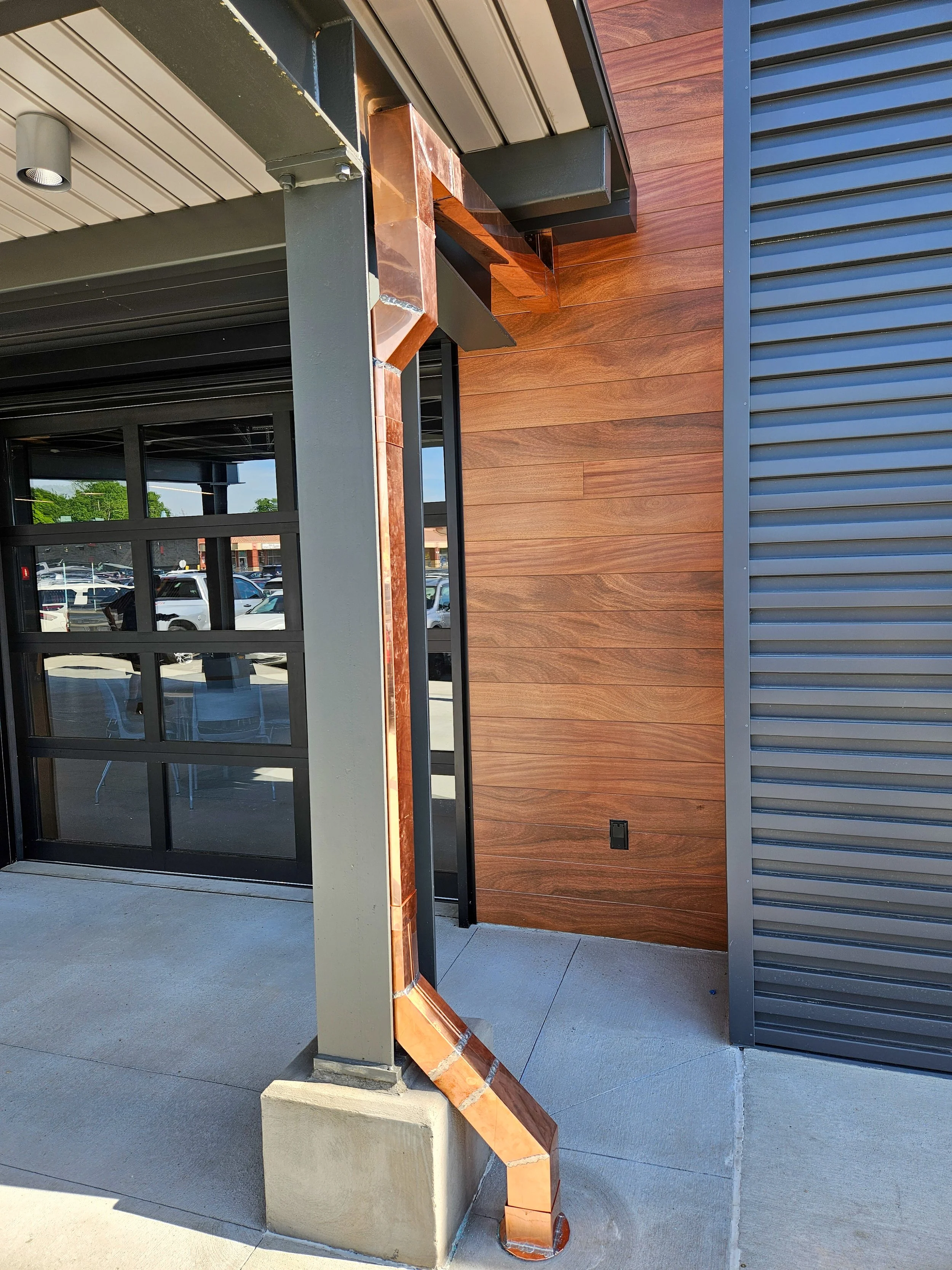 Exterior corner of a modern building showing a blue metal siding, wood paneling, and a copper downspout system with a concrete base.