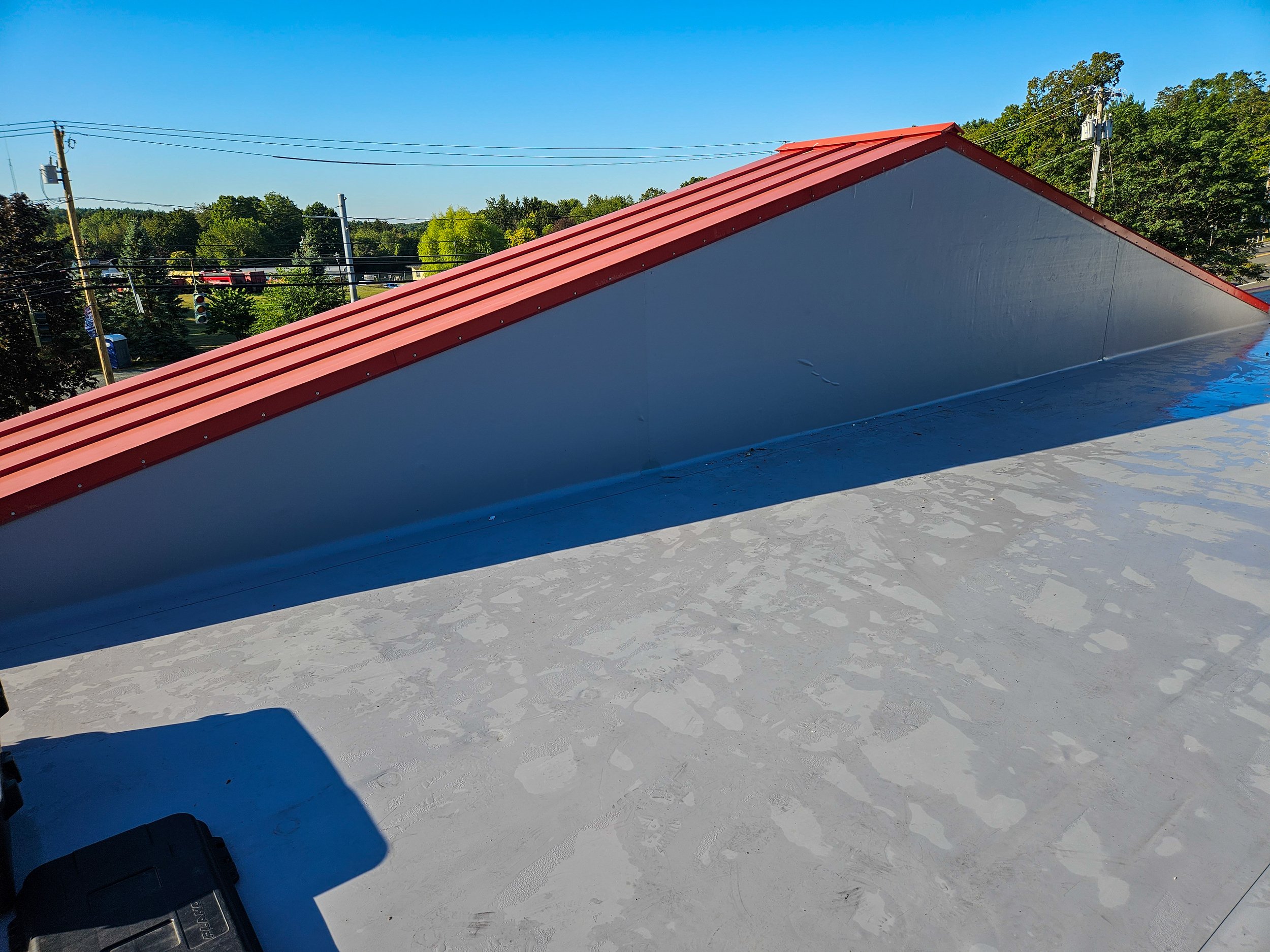 Roof with a sloped red metal panel and a flat gray section beneath, clear blue sky, trees and power lines in the background.