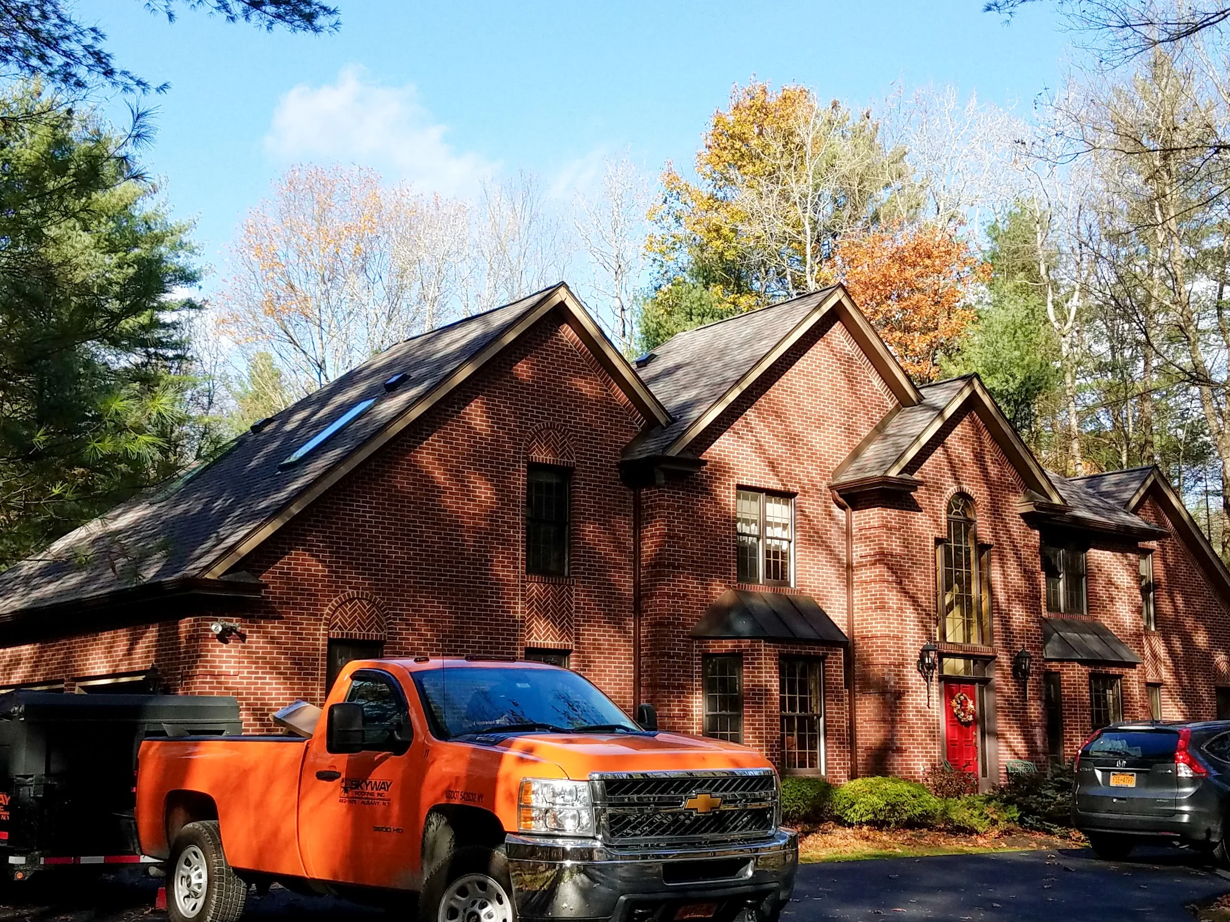A red brick house with multiple gable roofs, surrounded by trees with autumn foliage, and a bright blue sky in the background. There are two vehicles in the driveway, one is an orange Chevy truck and the other is a silver SUV. The house has tall, nar