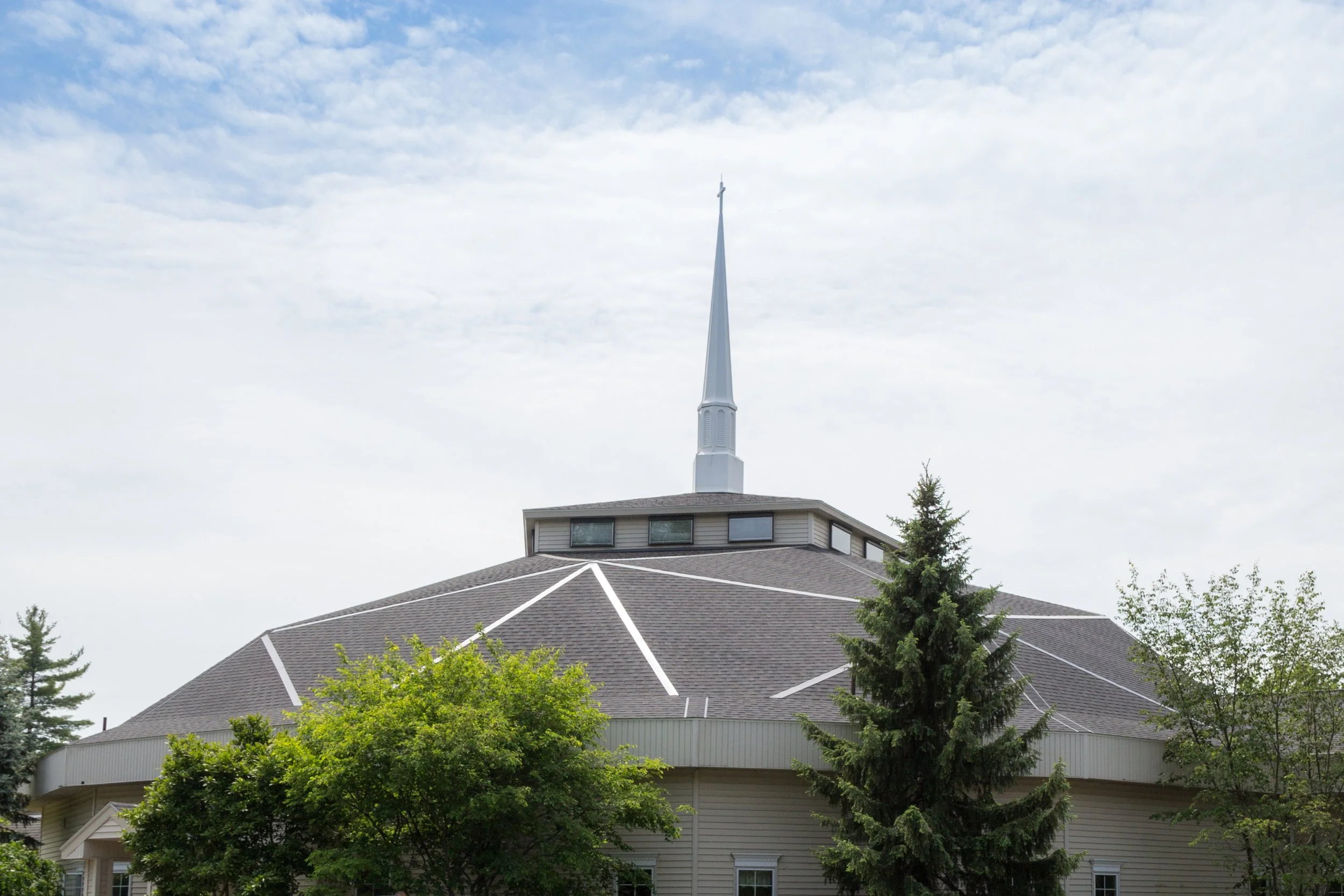 A church with a tall steeple surrounded by trees and partly cloudy sky.