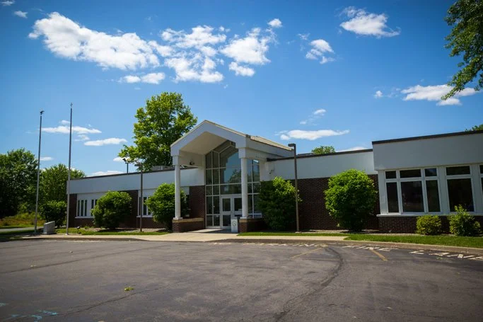 A single-story building with a modern entrance, surrounded by greenery, under a partly cloudy blue sky, with a parking lot in the foreground.