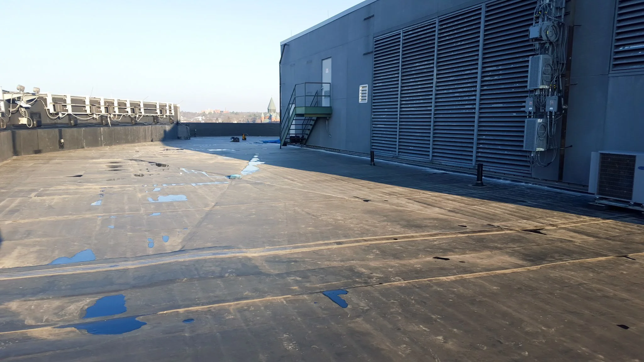 Rooftop surface with patches of water, side of a building with utility equipment, and a city skyline in the background.