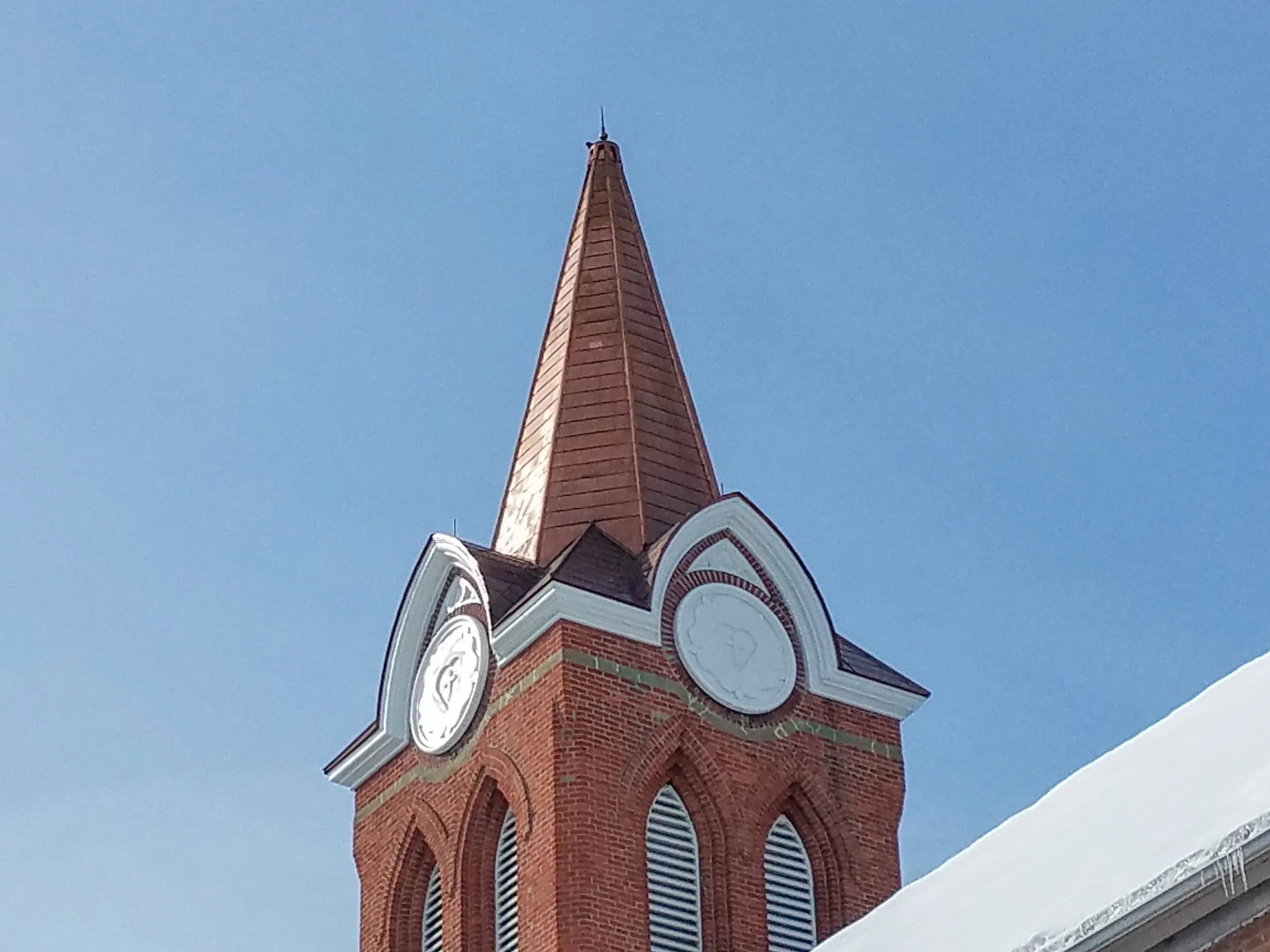 Close-up of a red brick church steeple with a clock, set against a clear blue sky, with snow at the bottom of the image.