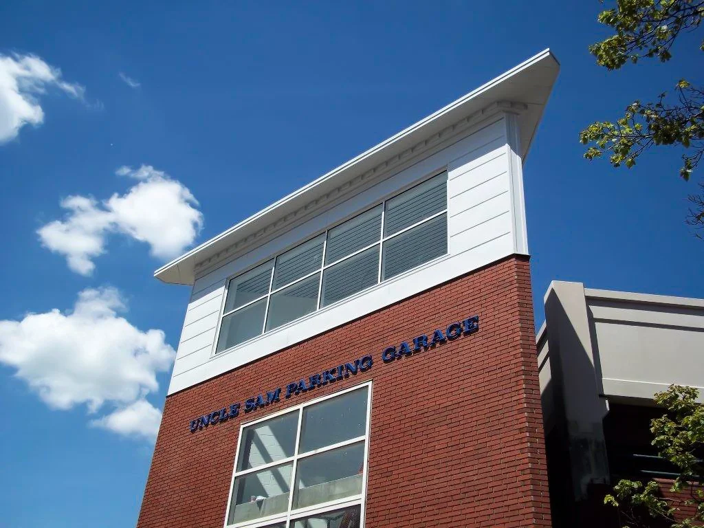 A multi-story building with a red brick facade and white siding, labeled 'Uncle Sam Parking Garage' in blue letters, against a partly cloudy blue sky.
