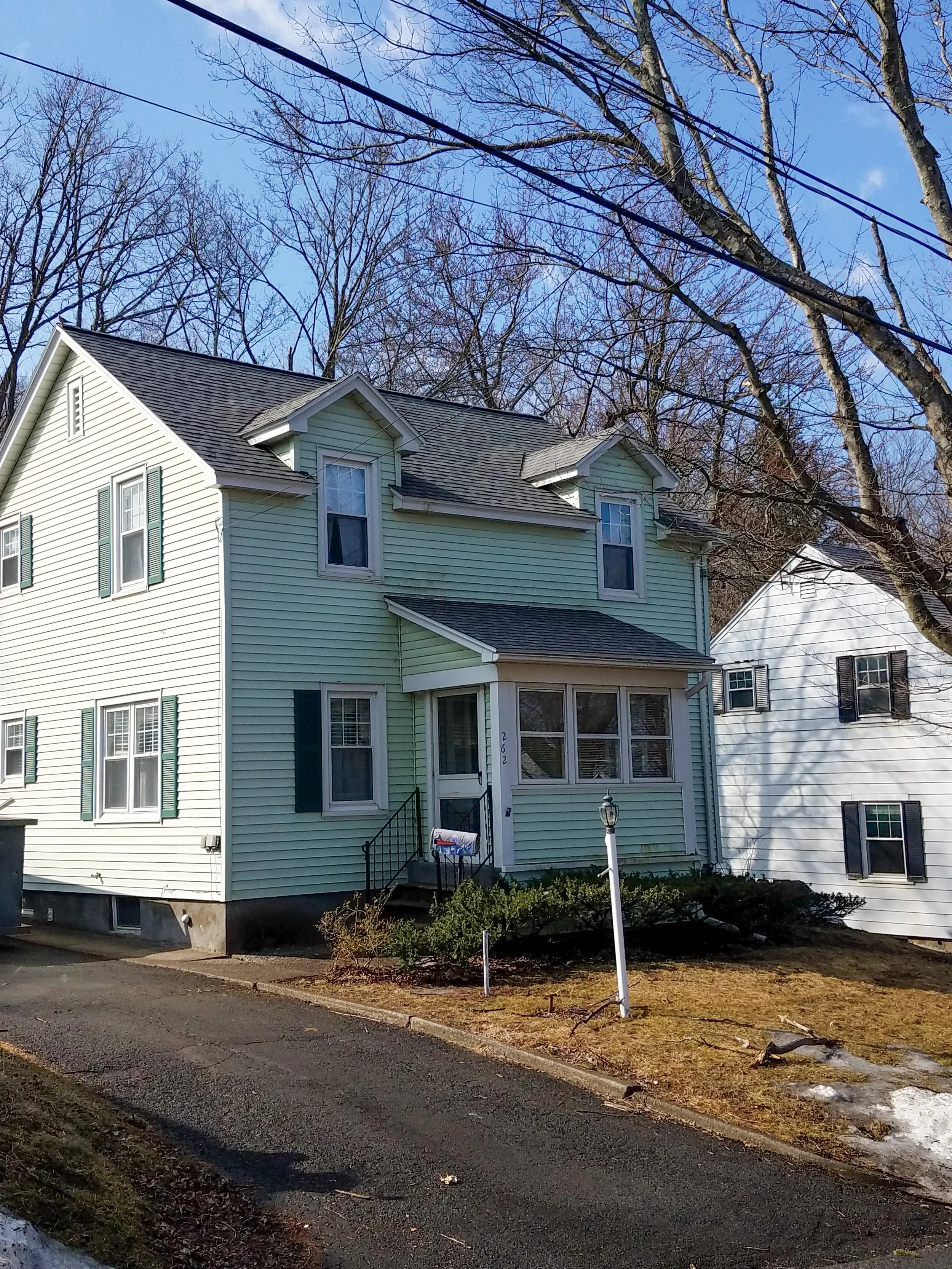 A two-story light green house with white trim and black shutters, with a porch and steps, surrounded by leafless trees and neighboring white houses, under a blue sky with some clouds.