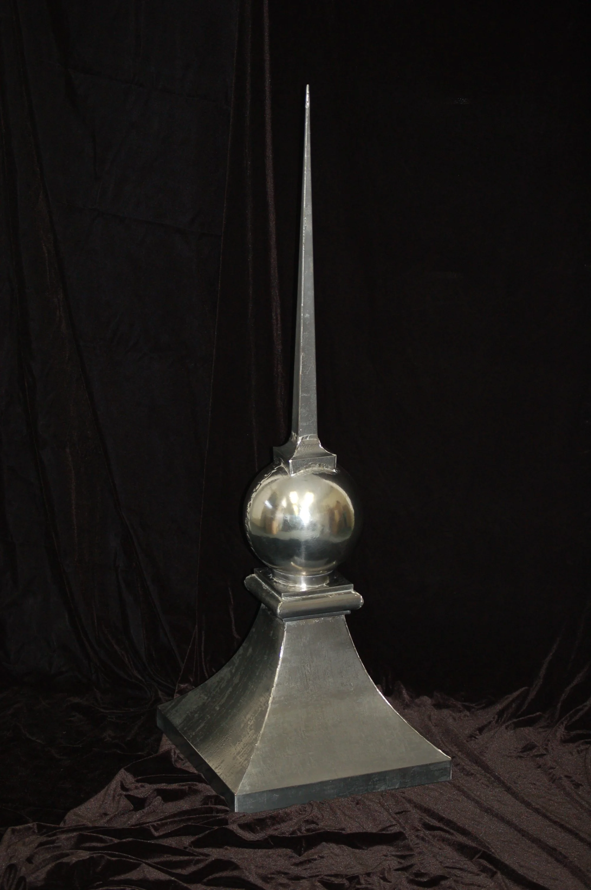A metallic sculpture featuring a large sphere with a long pointed spike on top, mounted on a wide, angled rectangular base.