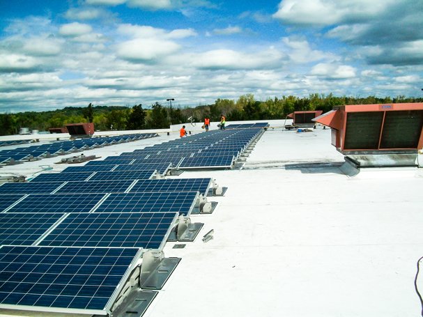 Photovoltaic solar panels installed on a flat commercial rooftop with workers in the background and a partly cloudy sky.