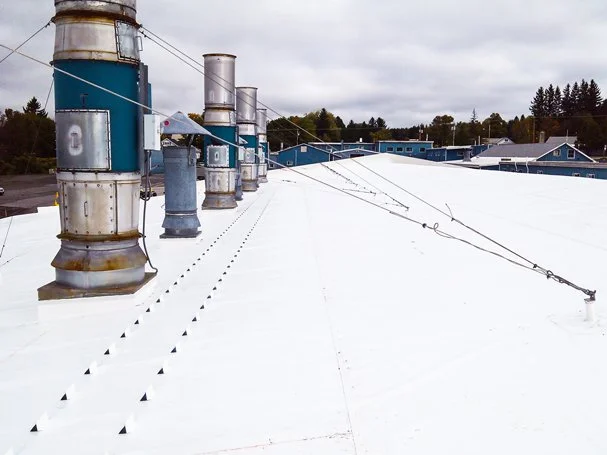 Industrial rooftop with multiple metal chimneys and vent pipes, some rusted, on a white surface under a cloudy sky.