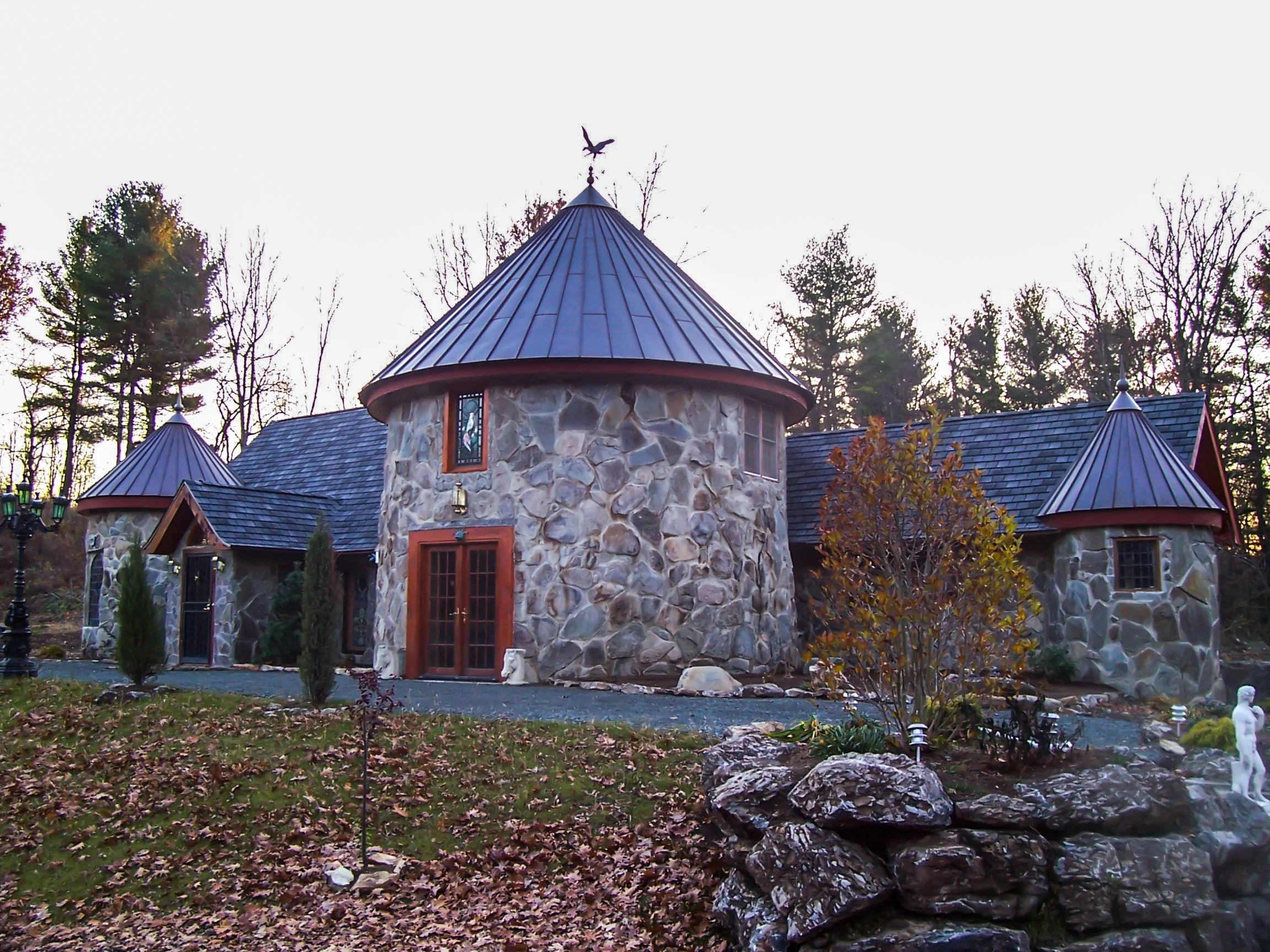 A stone house with a round tower and conical metal roof, surrounded by trees and fallen leaves.