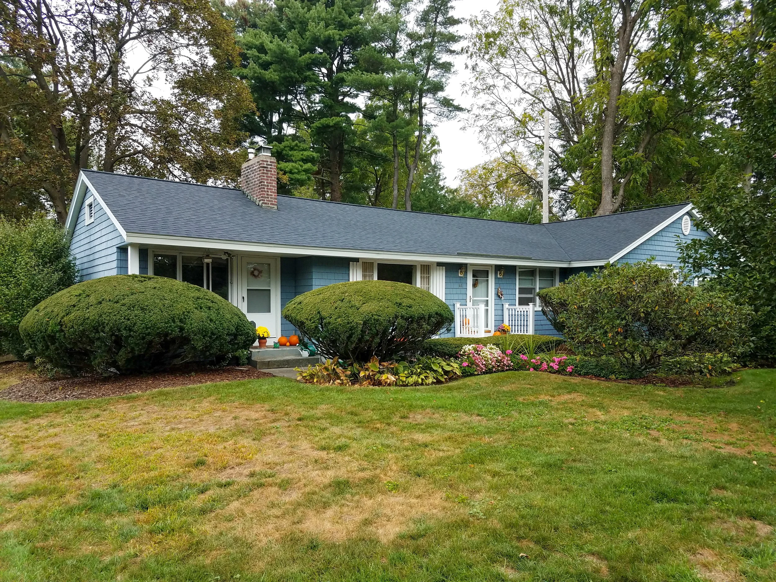 A blue single-story house with white trim and a dark gray roof, surrounded by well-kept shrubs and a lawn, with flowers and autumn decorations near the entrance.